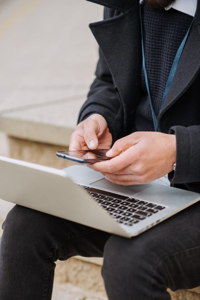 Crop anonymous male employee browsing internet on cellphone while sitting with netbook on staircase in city
