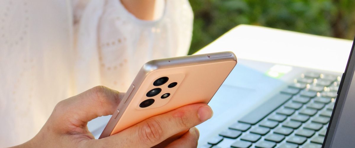 Person using a smartphone while drinking coffee near a laptop outdoors, showcasing multitasking with technology.